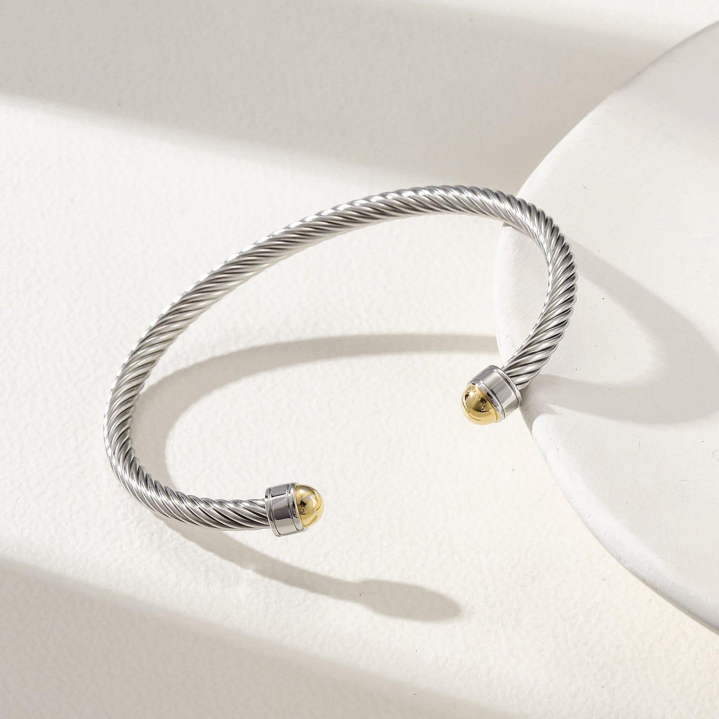 Silver twisted stainless steel cable cuff bracelet with 18k gold-plated end caps resting on a white display tray.