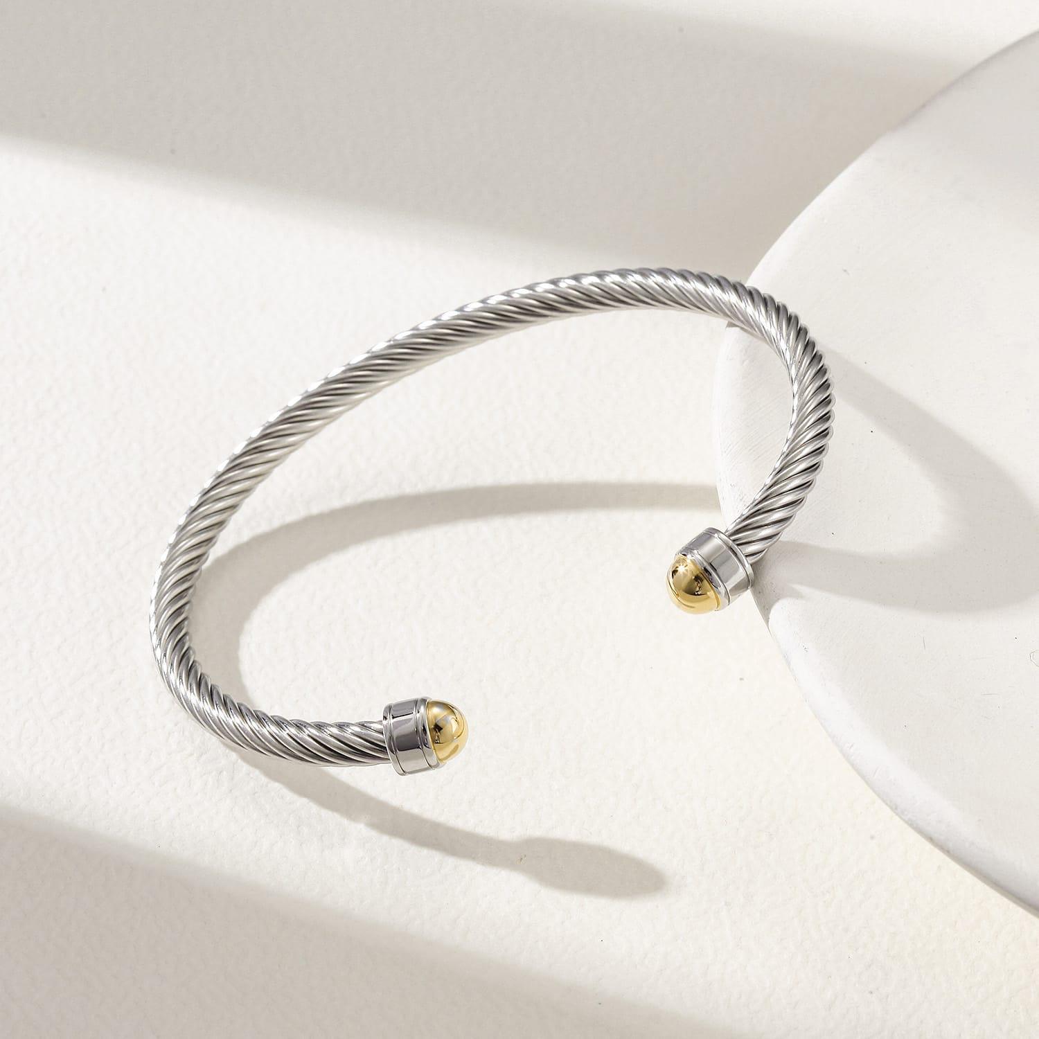 Silver twisted stainless steel cable cuff bracelet with 18k gold-plated end caps resting on a white display tray.